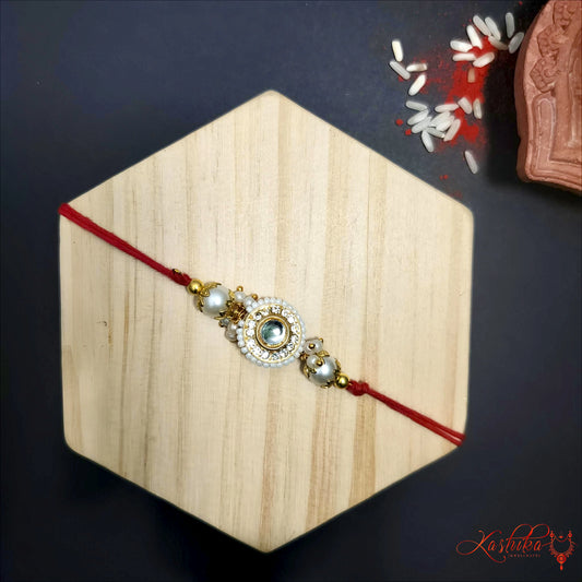 Elegant rakhi featuring Kundan style white stones and small pearls surrounding a central design , with gold tone accents and classic red thread against a subtle textured background