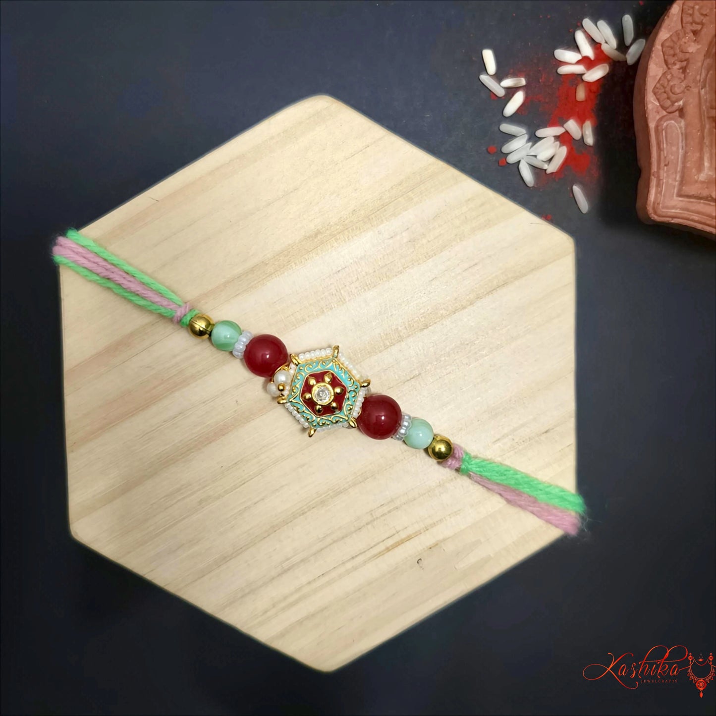 A single rakhi with green and pink threads , featuring a central ornate gold and white charm , flanked by red and light green beads , on a light wooden surface.
