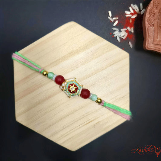 A single rakhi with green and pink threads , featuring a central ornate gold and white charm , flanked by red and light green beads , on a light wooden surface.