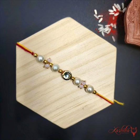  A single rakhi featuring a central Kundan stone , surrounded by white earls ( moti ) and small baby pink beads , strung on a yellow and red thread.