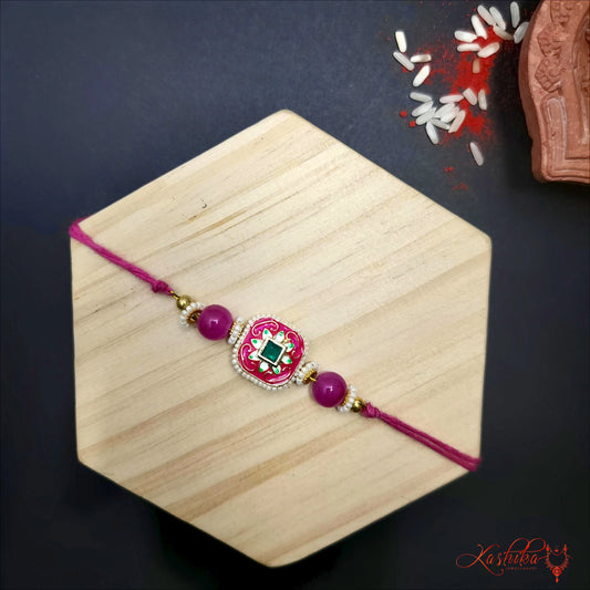 Pink rakhi with square enamel centerpiece , intricate Meenakari design and a greeen stonr , on a light wooden background