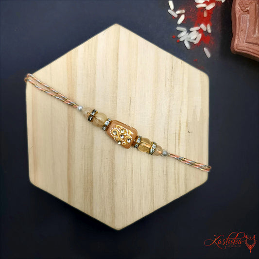 Light brown rakhi with an elongated centerpiece featuring small kundan stone work , flanked by brown and silver beads , on a light wooden background