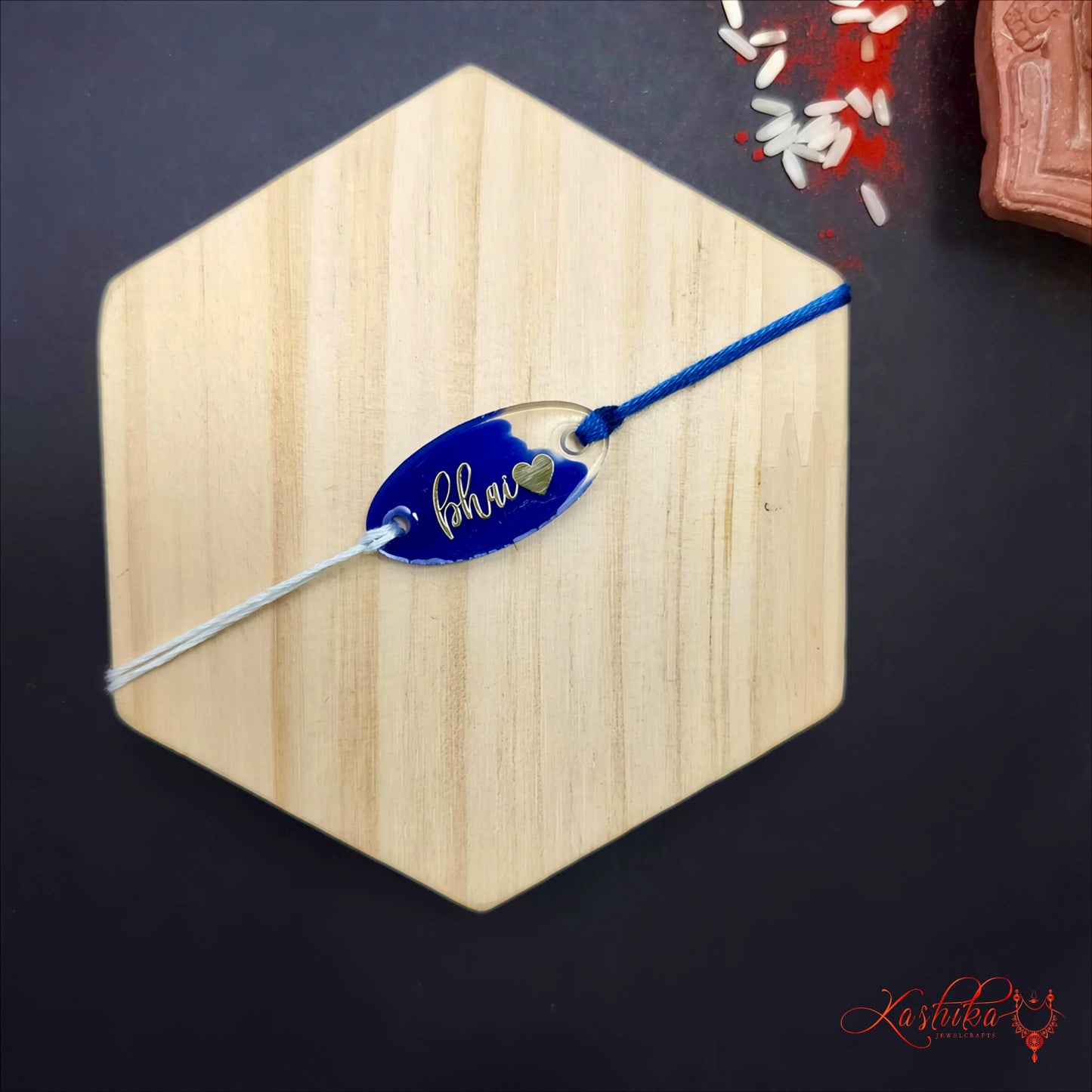 Blue Resin Rakhi with "Bhai" written in gold script and a small heart , clear oval charm , blue and white thread , on a light wooden surface