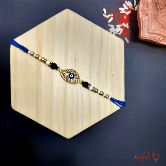 Evil Eye rakhi with blue and white design , surrounded by small golden stones, flanked by black and white beads, on a light wooden background