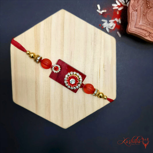 Dark red rakhi with rectangular centerpiece featuring a white floral design , red and golden beads