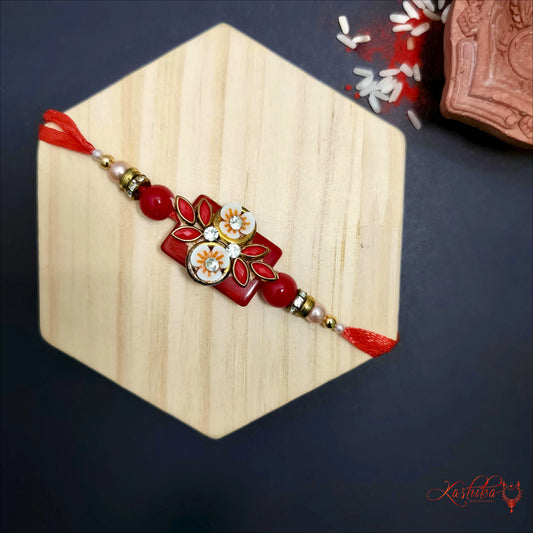 Dark red rakhi with a central rectangular design featuring white 3D flowers and red beads , on a wooden surface