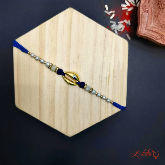 Rakhi with a golden hell evil eye charm at the center , flanked by white , blue and gold beads on a wooden board.