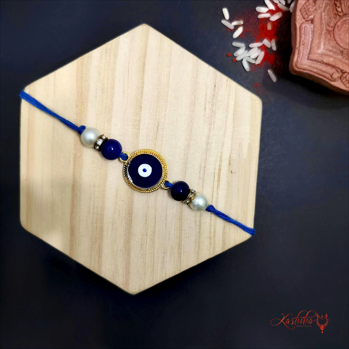 Blue rakhi  with a round golden-framed evil eye charm at center , flanked by blue beads and white pearls on wooden board