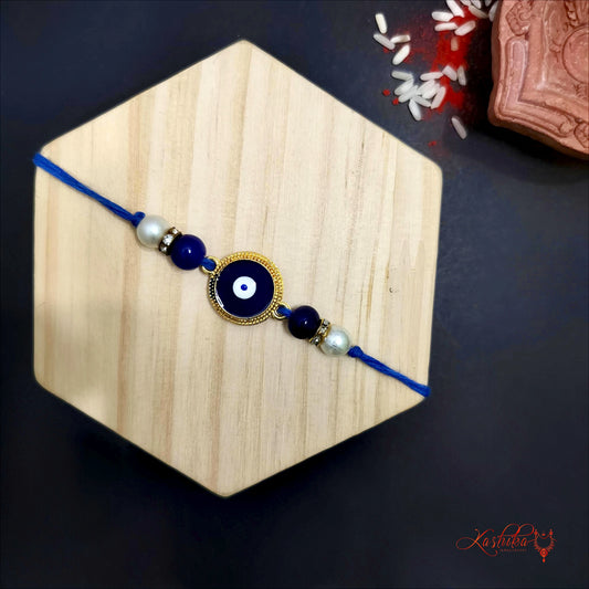 Blue rakhi  with a round golden-framed evil eye charm at center , flanked by blue beads and white pearls on wooden board