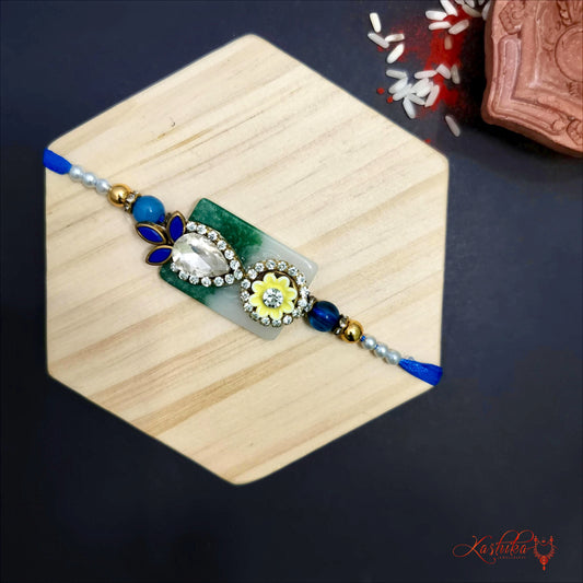 Blue and white rakhi with tear drop white stones ,blue petal shapes and a central golden floral design on a wooden hexagon