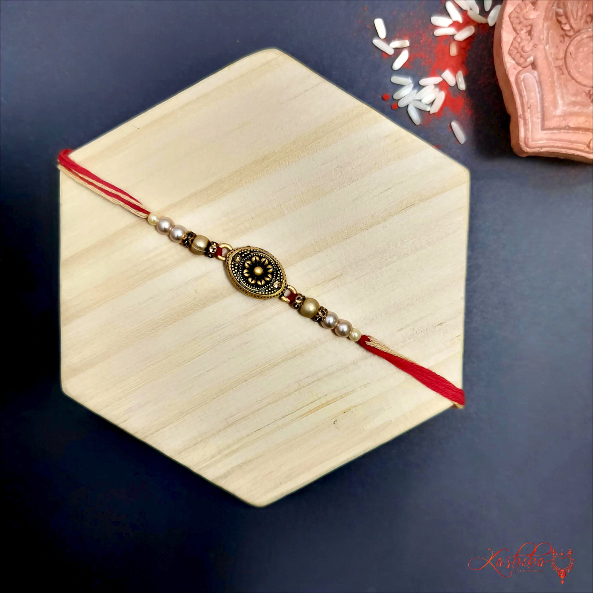 Red rakhi with a oval antique copper gold finish medallion at the center , flanked y golden and pearl - like beads on a wooden surface